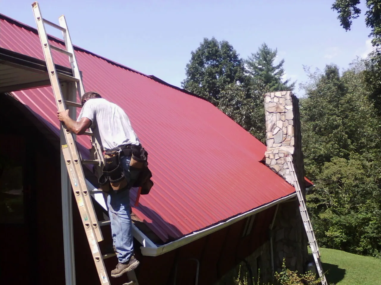 Roofer on a ladder beside a red metal roof and stone chimney — Soffit & Fascia Repair by Roofing Brothers in Bordentown
